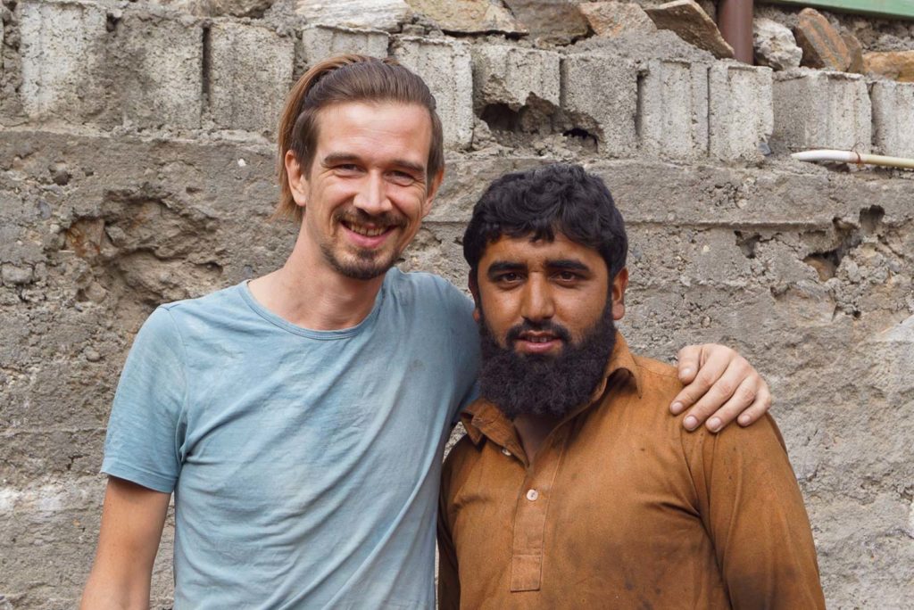 Ein Mann aus Pakistan und ein Mann aus Deutschland umarmen sich und lachen in die Kamera. eine Steiunwand ist im Hintergrund.