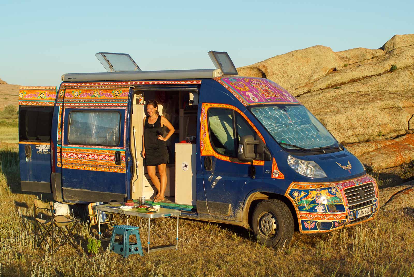 Frau steht in einem geöffneten Camperbus und Blick hinaus. Sie blickt in den Sonennschein. Der Camper steht in der Natur , der steppe. Vor der Camper eine gedeckter Essenstisch.