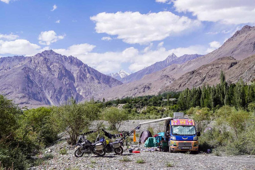 pakistans norden tief im karakorumgebirge. steinklolosse ragen gegen himmel. himmel mit weisse wolken. zwei motoräder im vordergrund und ein bunter kastenwagen. die markise ist ausgefahren und eine grünes zelt steht daneben.