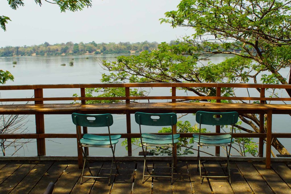 drei grüne stühle stehen in thailand vor dem mekong auf einem holzpodest. seitlich raken große grüne bäume ins bild. mekong ist sehr ruhig
