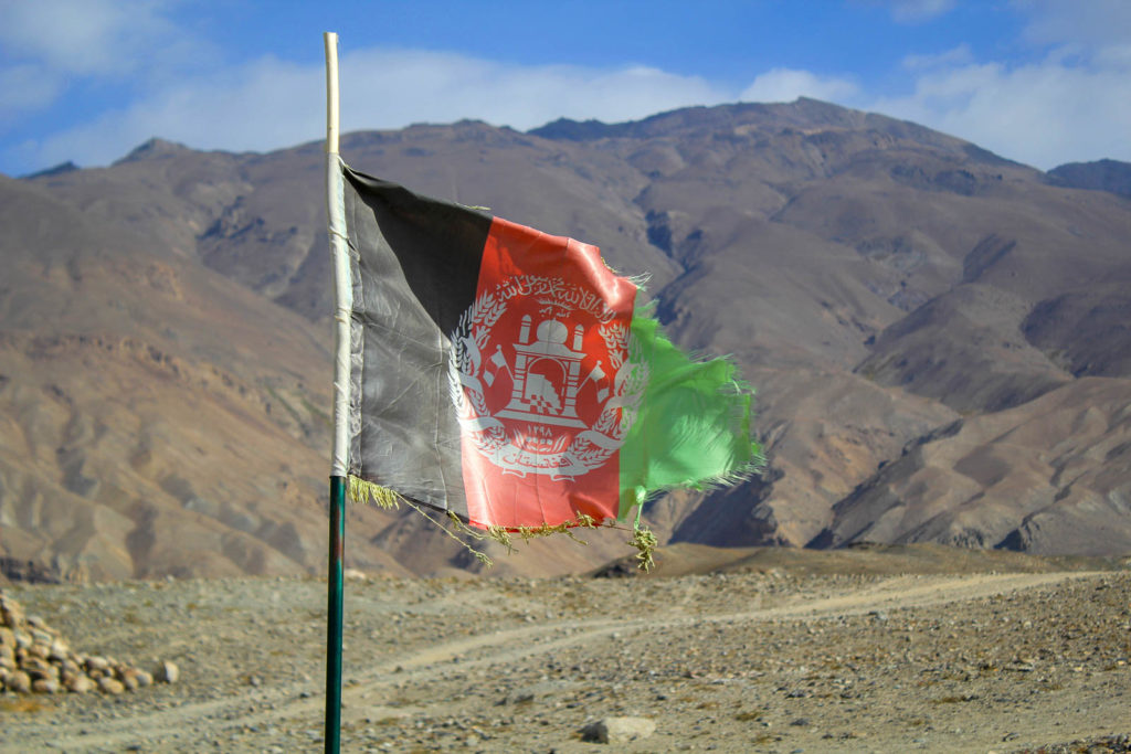 in die jahre gekommene afganische flagge weht im wind. sie ist gestreift schwarz rot grün und eine wenig angerissen. im hintergrund bergigie landschaft und blauer himmel