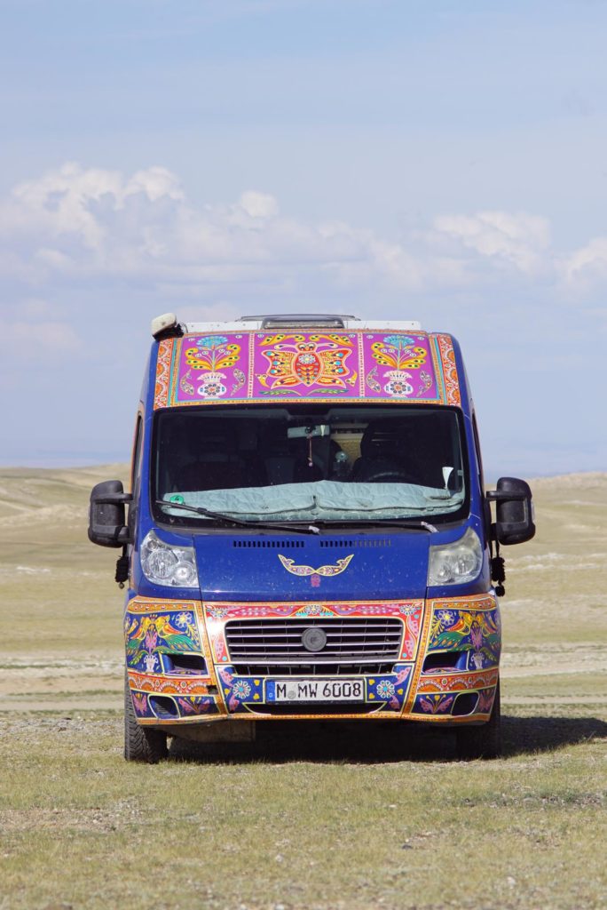 Das bunter selbstausgebaute Wohnmobil steht mitten in der Steppenlandschaft der Mongolei. In Hintergrund scheint die sonne.